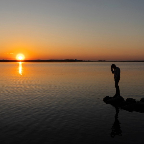 Man holding head sadly while looking at the water at sunset